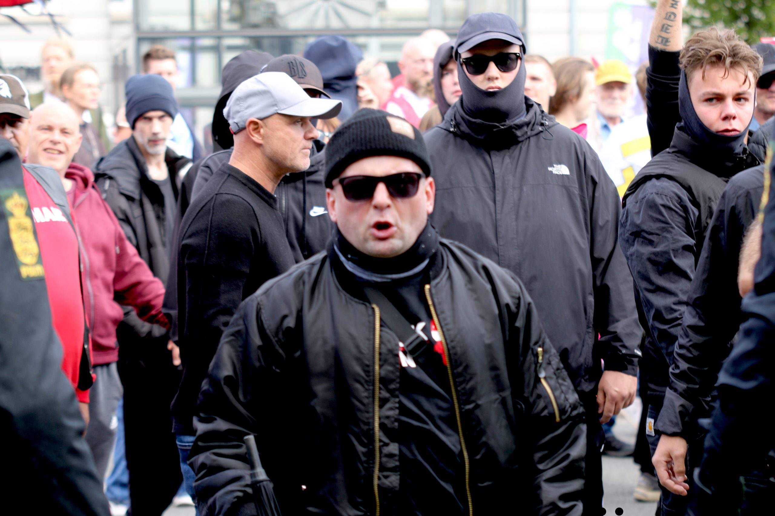 Midt i billedet ses den gamle Brøndby-hooligan André Stilund Vincent. Lige bag ham, i profil med lys kasket, ses Brian ‘Cat’ Jensen, der ligesom Vincent var en del af det nynazistiske netværk Dansk Front. Yderst til højre uden hovedbeklædning ses Frederik Munk, der udover at være en del af Active Club Denmark er en del af det uofficielle fanmiljø omkring FC København.